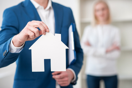 Close up of cheerful realtor selling an apartment. He is holding a placard in the shape of house. The woman is standing on background